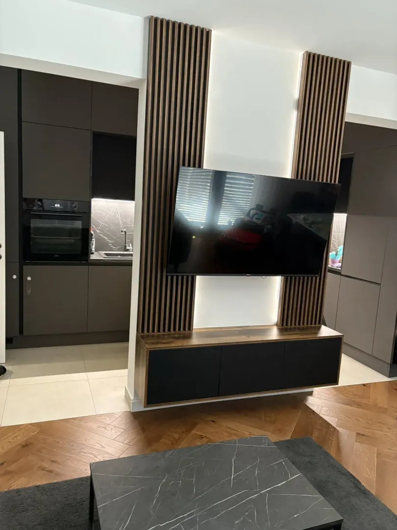 Moderni dnevni boravak sa TV policom i kuhinjom u pozadini.