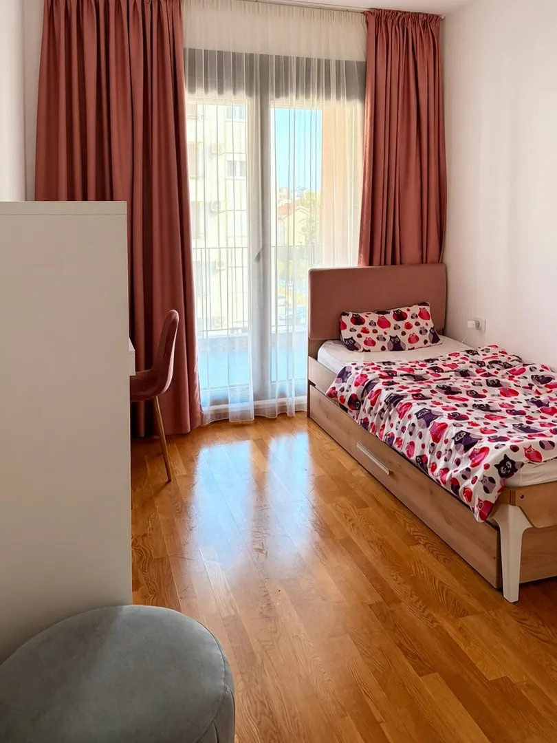 Manja spavaća soba sa krevetom, radnim stolom i prozorom sa roze zavjesama.
