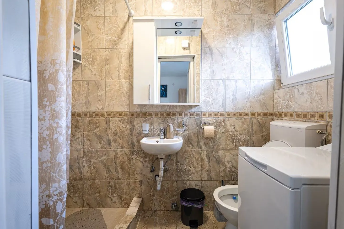 Manje kupatilo sa umivaonikom, ogledalom i WC šoljom obloženo keramičkim pločicama.