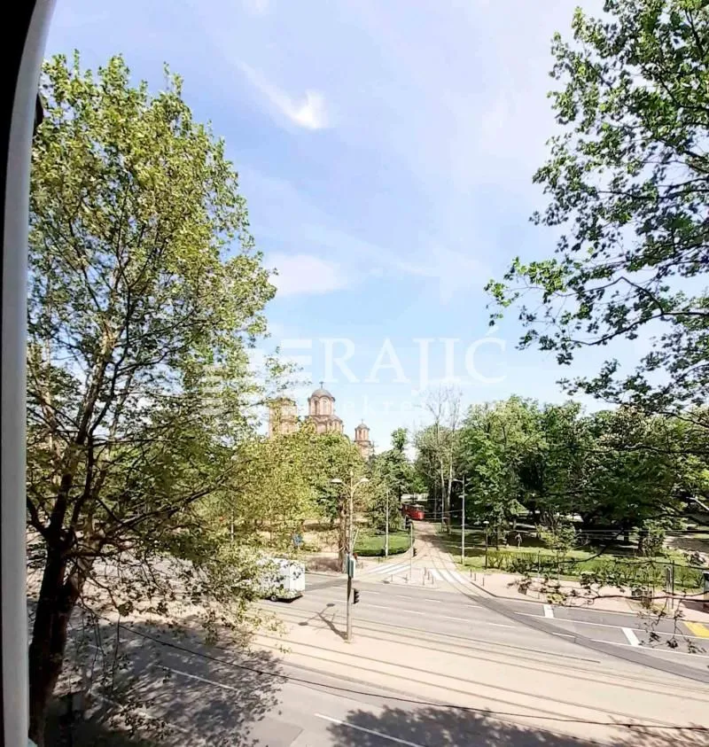 Pogled sa prozora na ulicu i park sa drvećem.