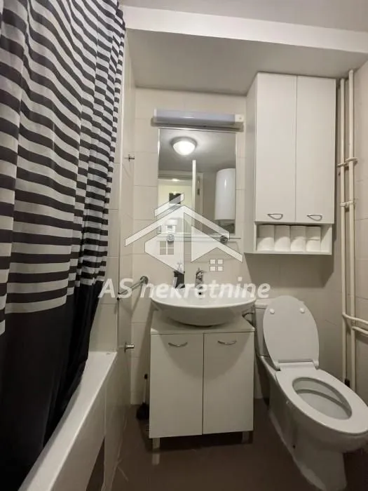 Manje kupatilo sa umivaonikom, WC šoljom, ormarićem i kadom sa zavesom.