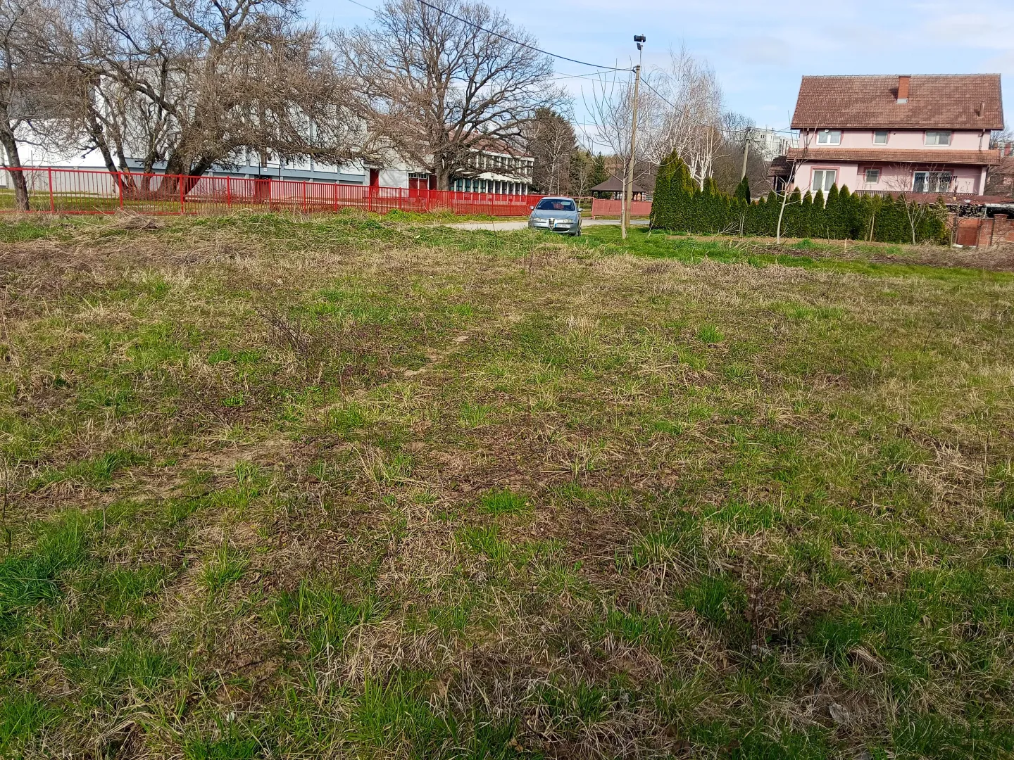 Pogled na prostrani travnati plac sa drvećem i okolnim kućama.