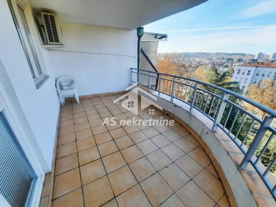 Prostrani popločani balkon sa metalnom ogradom i pogledom na naselje.