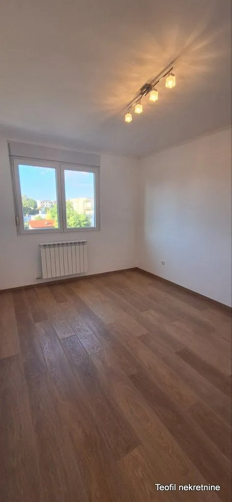 Prazna soba sa belim zidovima, drvenim podom, prozorom i radijatorom.