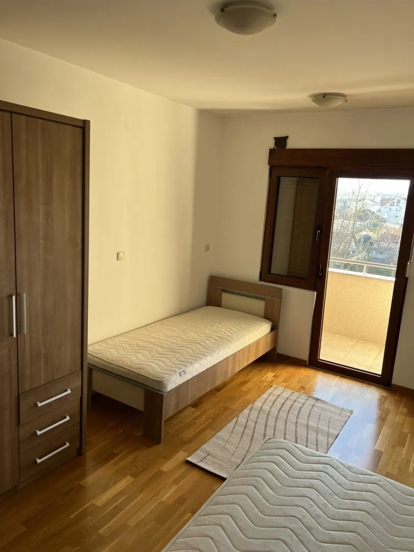 Jednostavna spavaća soba sa jednim krevetom, drvenim ormarom i izlazom na balkon.
