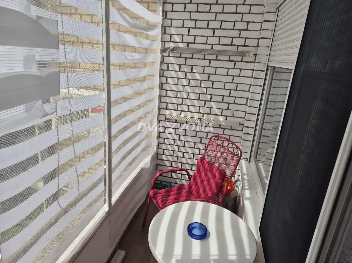 Zastakljeni balkon sa malim stolom, crvenom stolicom i belom ciglom na zidu.
