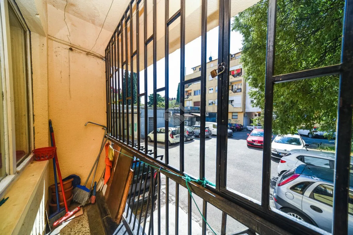 Manji natkriveni balkon sa metalnom ogradom i pogledom na parking i zgrade.