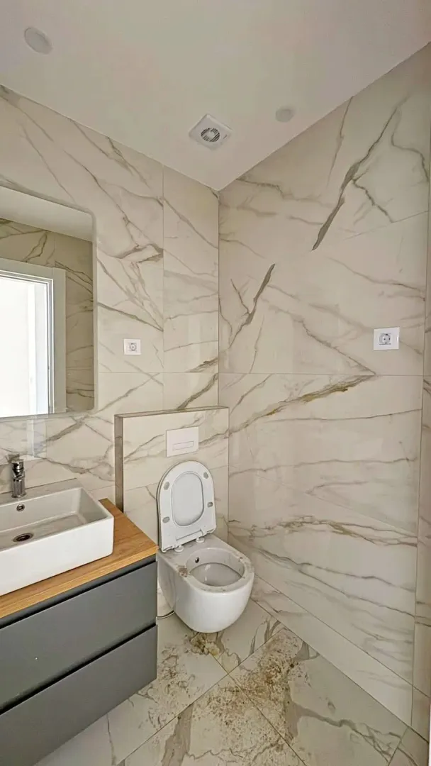 Modern bathroom with a sink, mirror, and toilet lined with marble tiles.