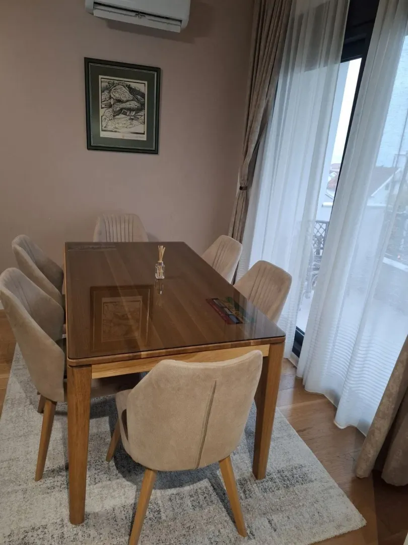 Trpezarija sa drvenim stolom i tapaciranim stolicama pored velikog prozora.