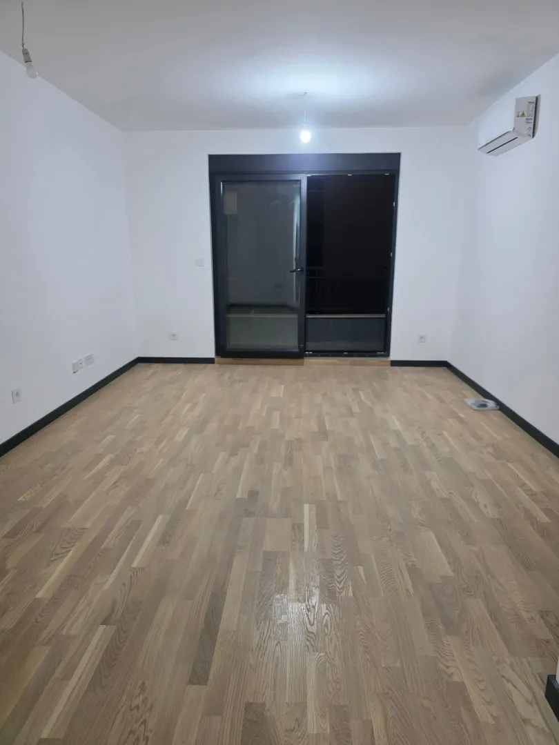 Prazna soba sa parketom, bijelim zidovima i klima-uređajem.
