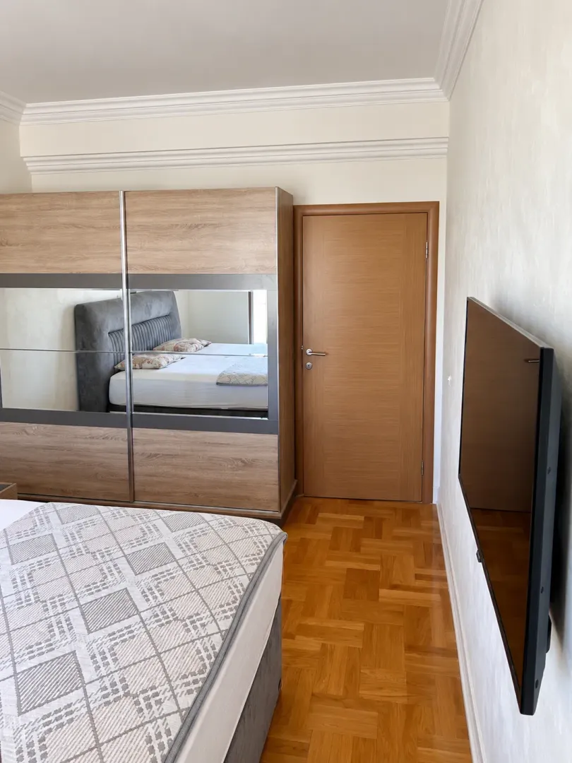 Spavaća soba sa bračnim krevetom, drvenim ormarom sa ogledalom i zidnim televizorom.