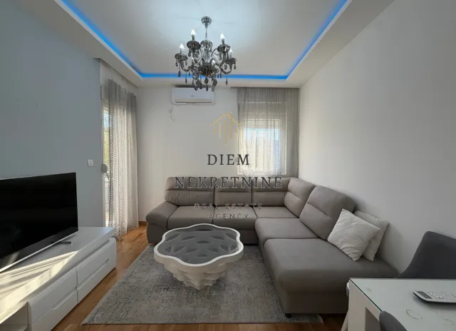 Dnevni boravak sa ugaonom garniturom, televizorom i trpezarijskim stolom.