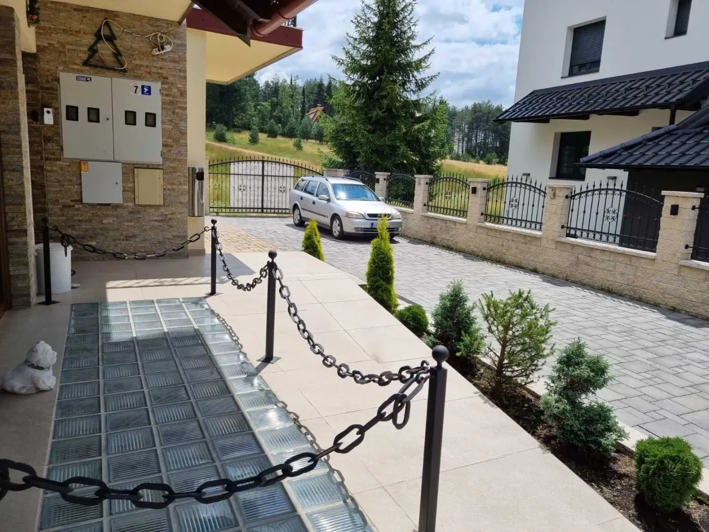 Eksterijer kuće sa popločanim dvorištem, ogradom i parkiranim automobilom.