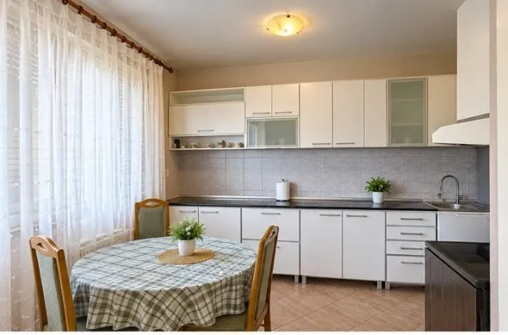 Bijela kuhinja sa trpezarijskim stolom i stolicama pored prozora.