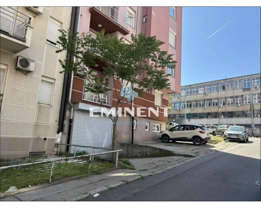 Pogled na spoljašnjost zgrade sa parkingom i drvetom u dvorištu.
