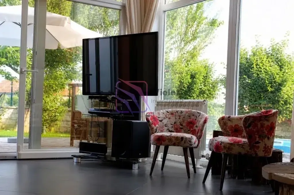 Living room with a TV, armchairs and large glass surfaces overlooking the yard.