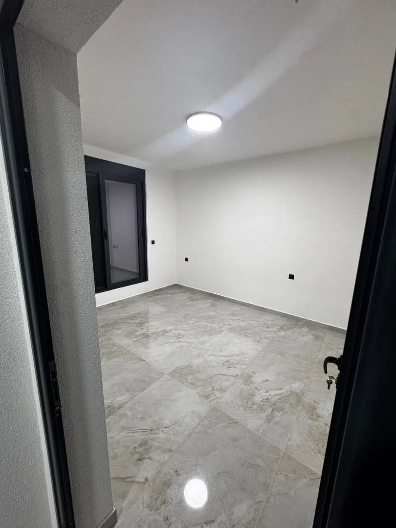 Prazna prostorija sa keramičkim pločicama i balkonskim vratima, viđena kroz vrata.