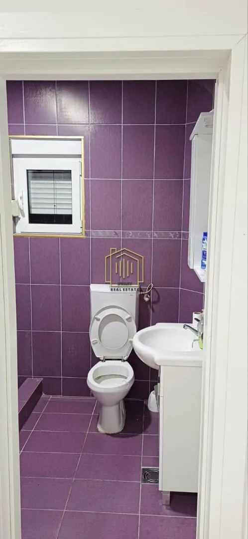 Kupatilo obloženo ljubičastim pločicama sa WC šoljom i umivaonikom.