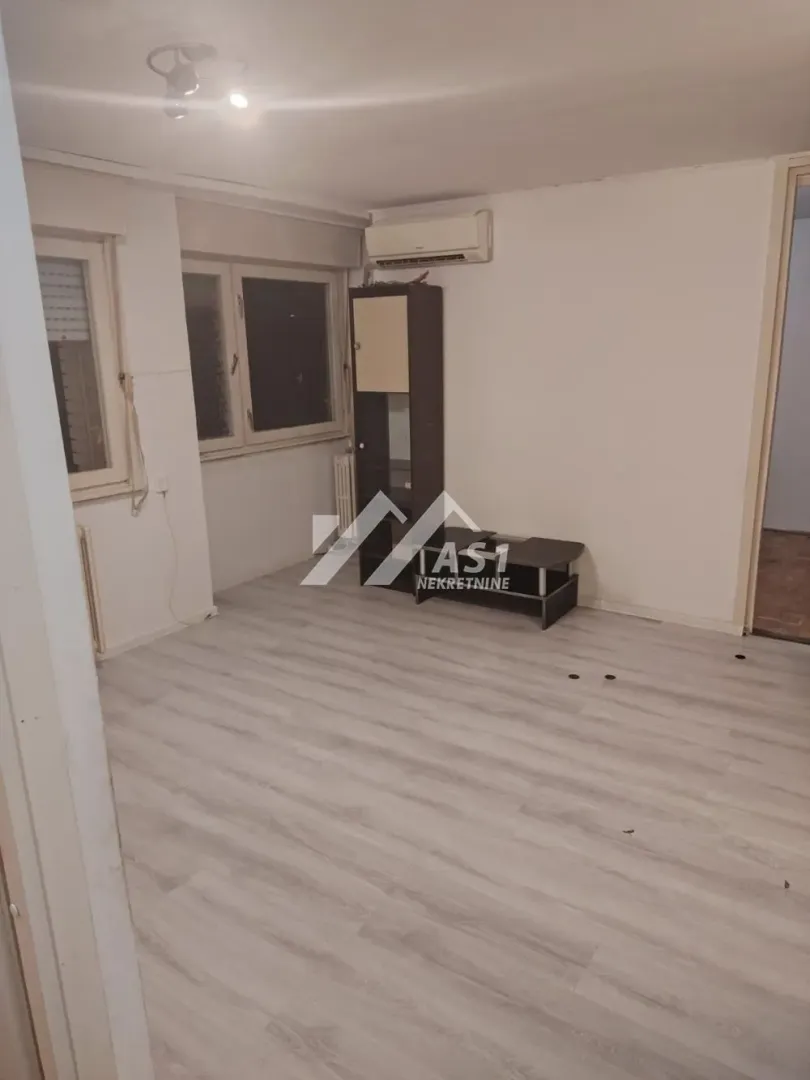 Prazna dnevna soba sa svetlim laminatom, klima uređajem i komadom nameštaja.