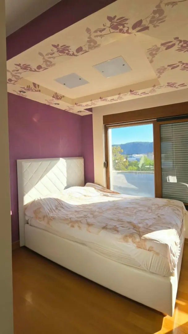 Spavaća soba sa bračnim krevetom, ljubičastim zidom i dekorativnim plafonom.