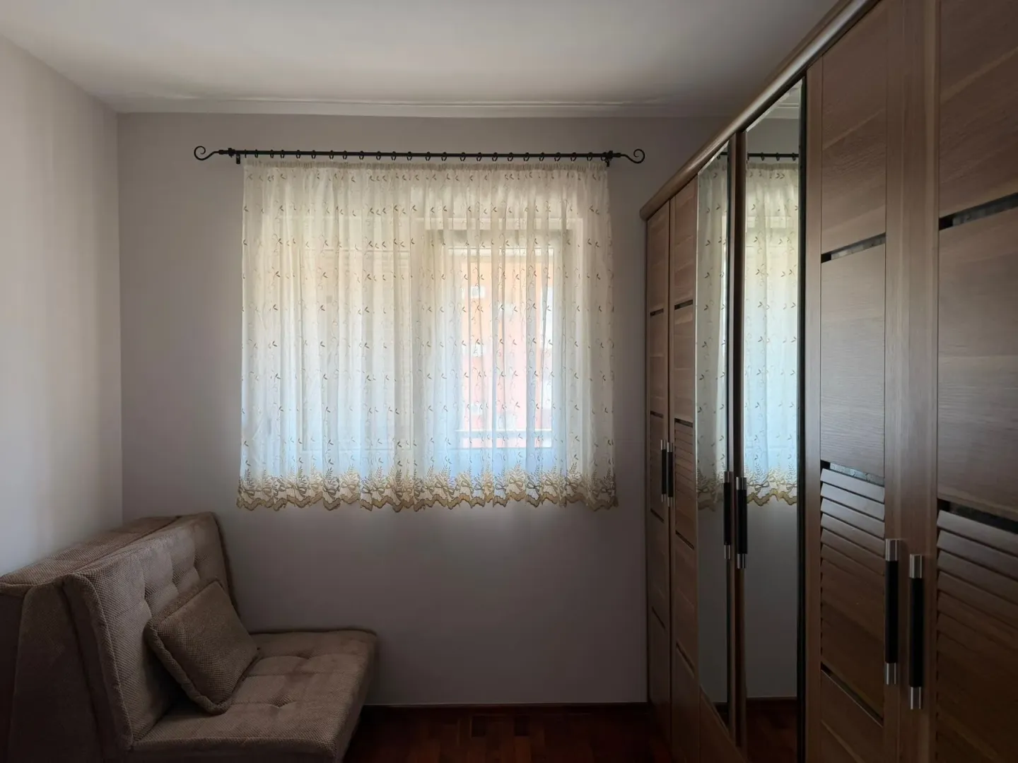 Small bedroom with a sofa bed, sheer curtains, and a large wooden wardrobe with a mirror.