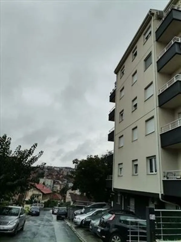 Pogled na spoljašnjost stambene zgrade i ulicu sa parkiranim automobilima.