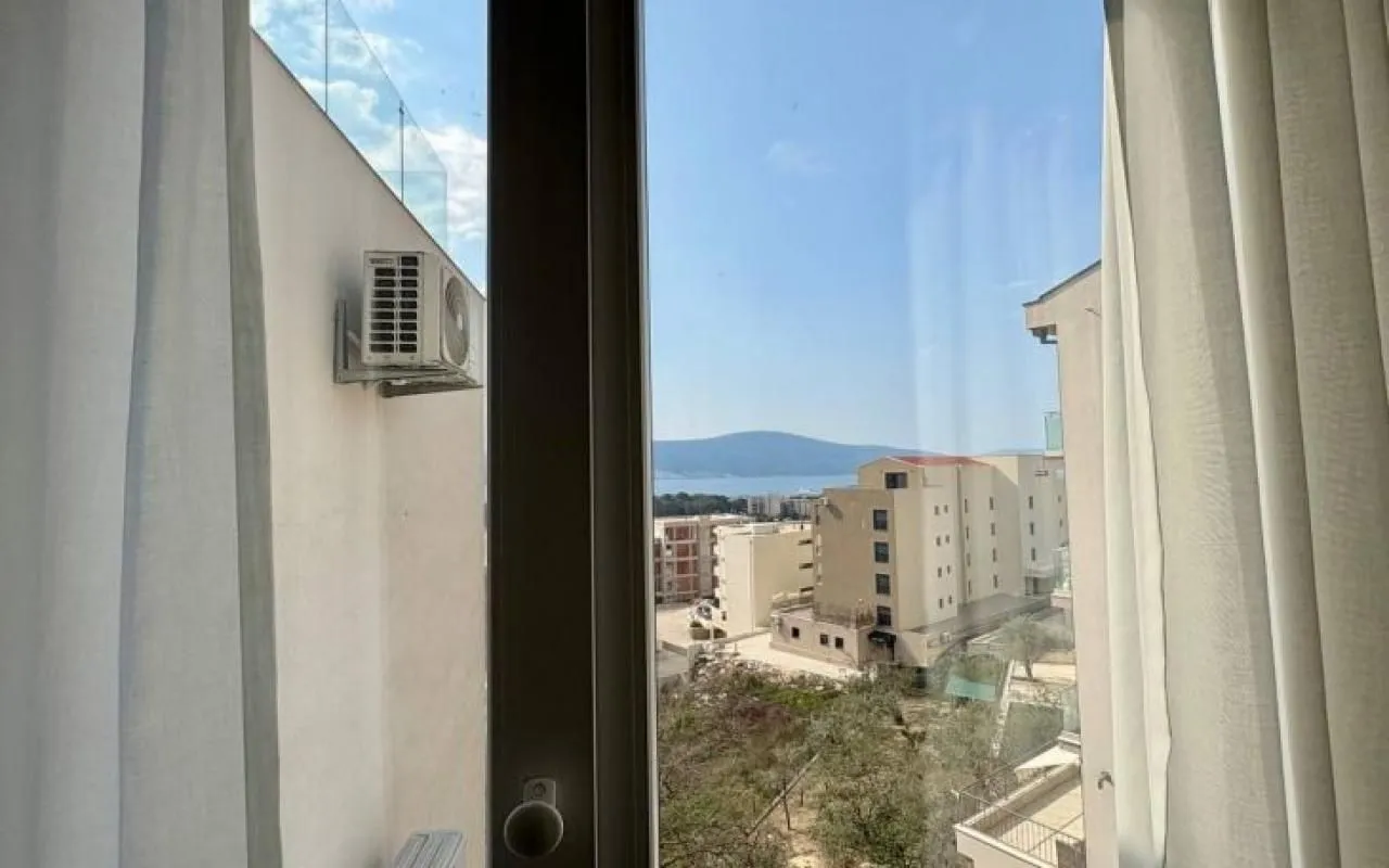 Pogled kroz prozor sa bijelim zavesama na okolne zgrade i more u daljini.