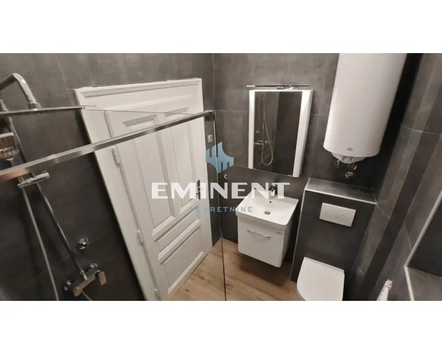 Moderno kupatilo obloženo tamnim pločicama sa tuš kabinom, umivaonikom i wc šoljom.