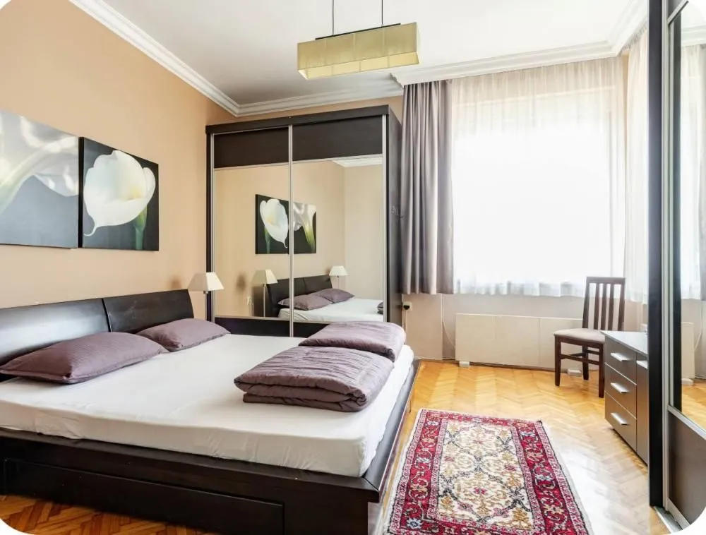 Furnished bedroom with a double bed, large mirrored wardrobe, and wooden flooring.