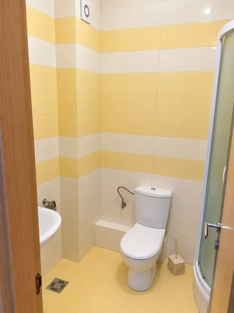 Manje kupatilo sa žuto-bijelim pločicama, wc šoljom, umivaonikom i tuš kabinom.