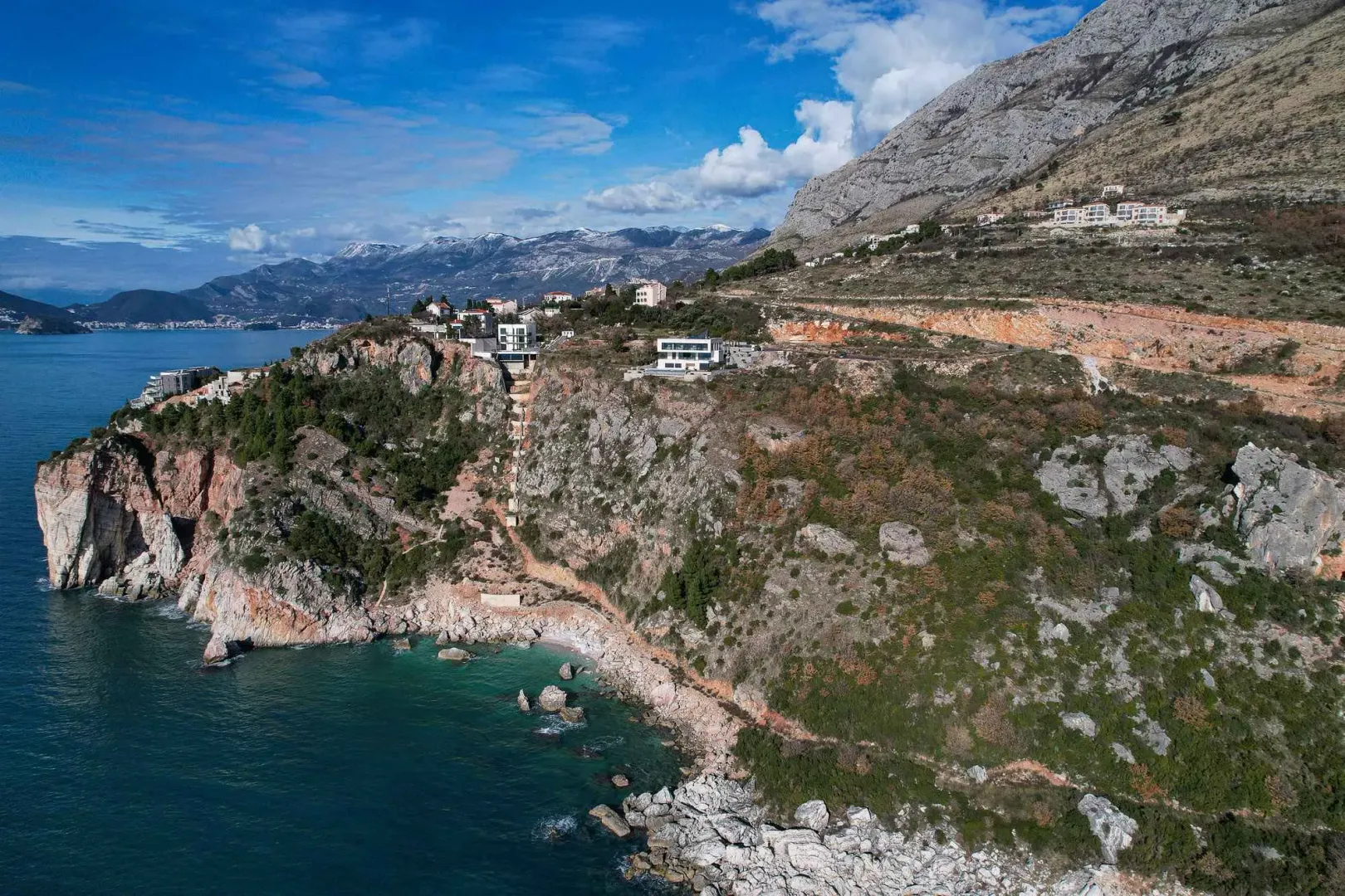 Pogled iz vazduha na obalu sa kućama na litici i planinama u pozadini.