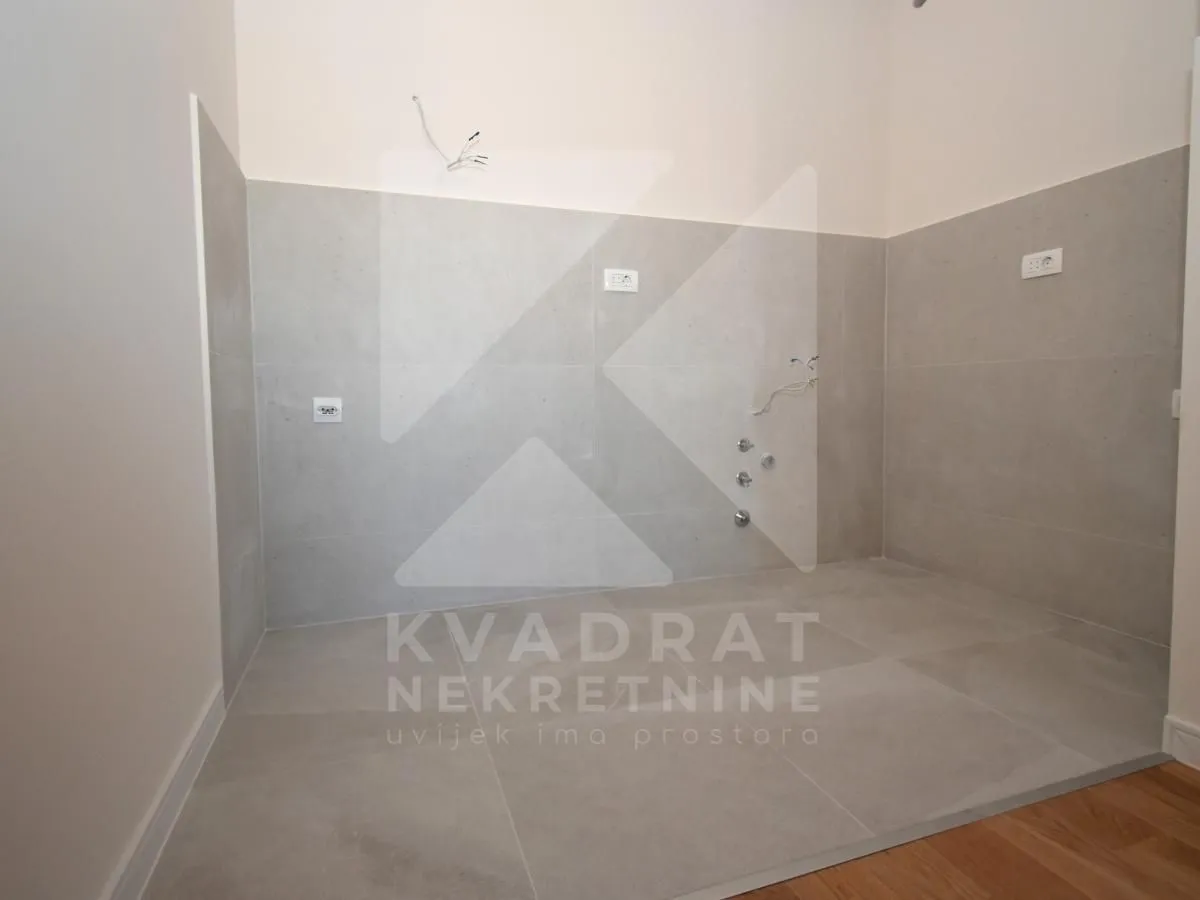 Nenamješten dio kuhinje sa sivim zidnim pločicama i pripremljenim priključcima za vodu i struju.
