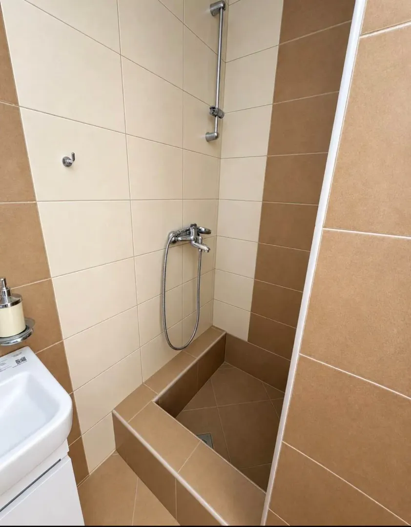 Prikaz kupatila sa tuš kabinom i keramičkim pločicama u bež i smeđim tonovima.