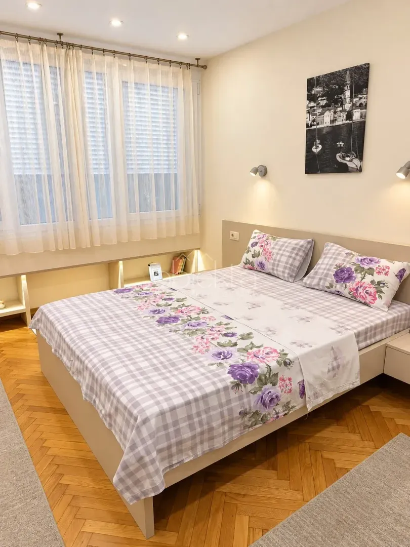 Furnished bedroom with a double bed, parquet flooring, and a large window.