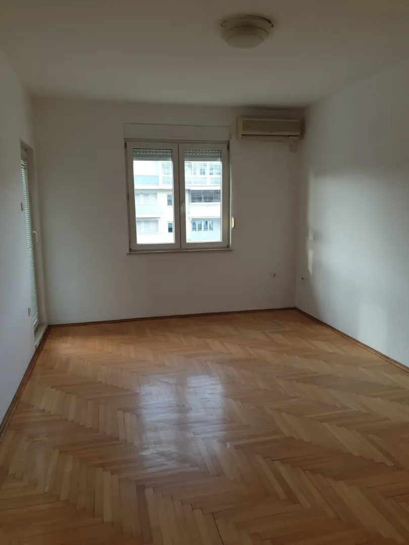 An empty room with parquet flooring, a window, and an air conditioner.