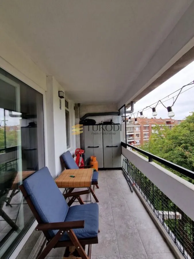 Covered balcony with a table, chairs, and a view of greenery and surrounding buildings.