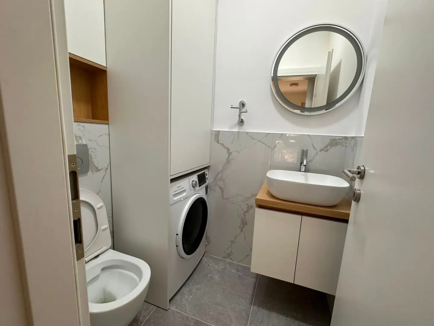 Moderne kupatilo sa umivaonikom, WC šoljom, mašinom za veš i okruglim ogledalom.