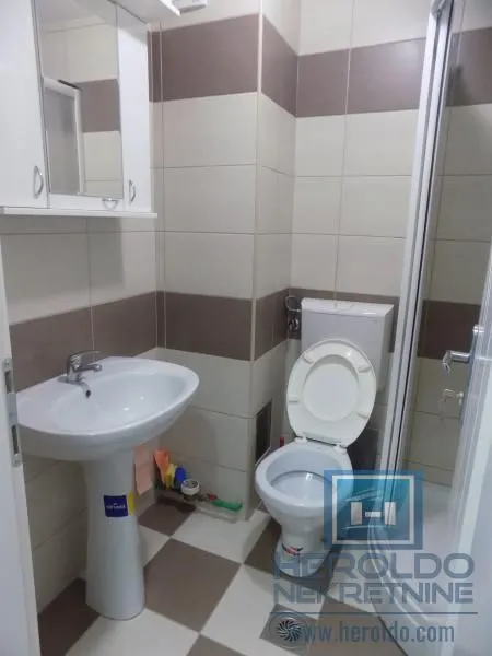 Manje kupatilo sa umivaonikom, WC šoljom i tuš kabinom obloženo keramičkim pločicama.