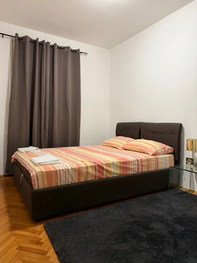 Furnished bedroom with a double bed, dark curtains, and parquet flooring.