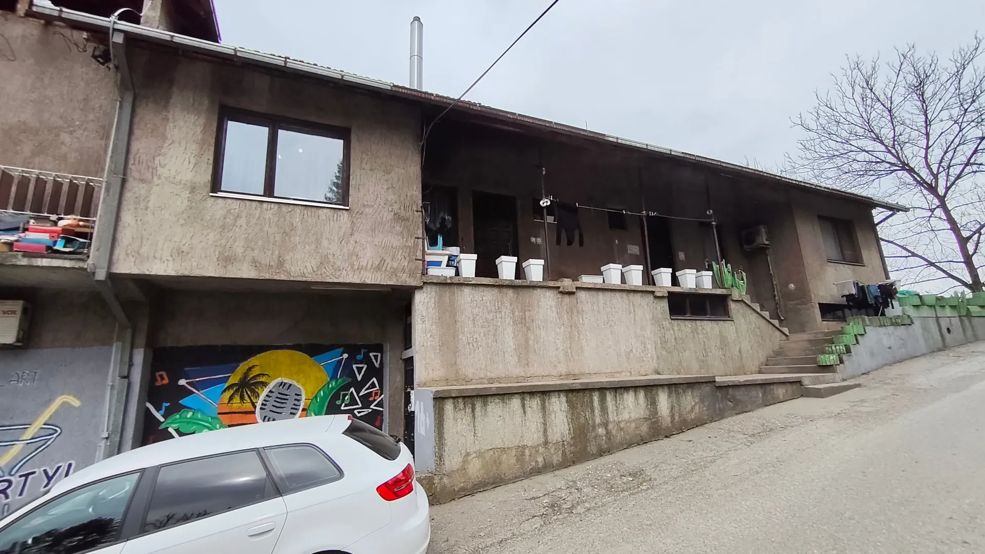Spoljašnji izgled starije kuće sa dvorištem i parkiranim automobilom.