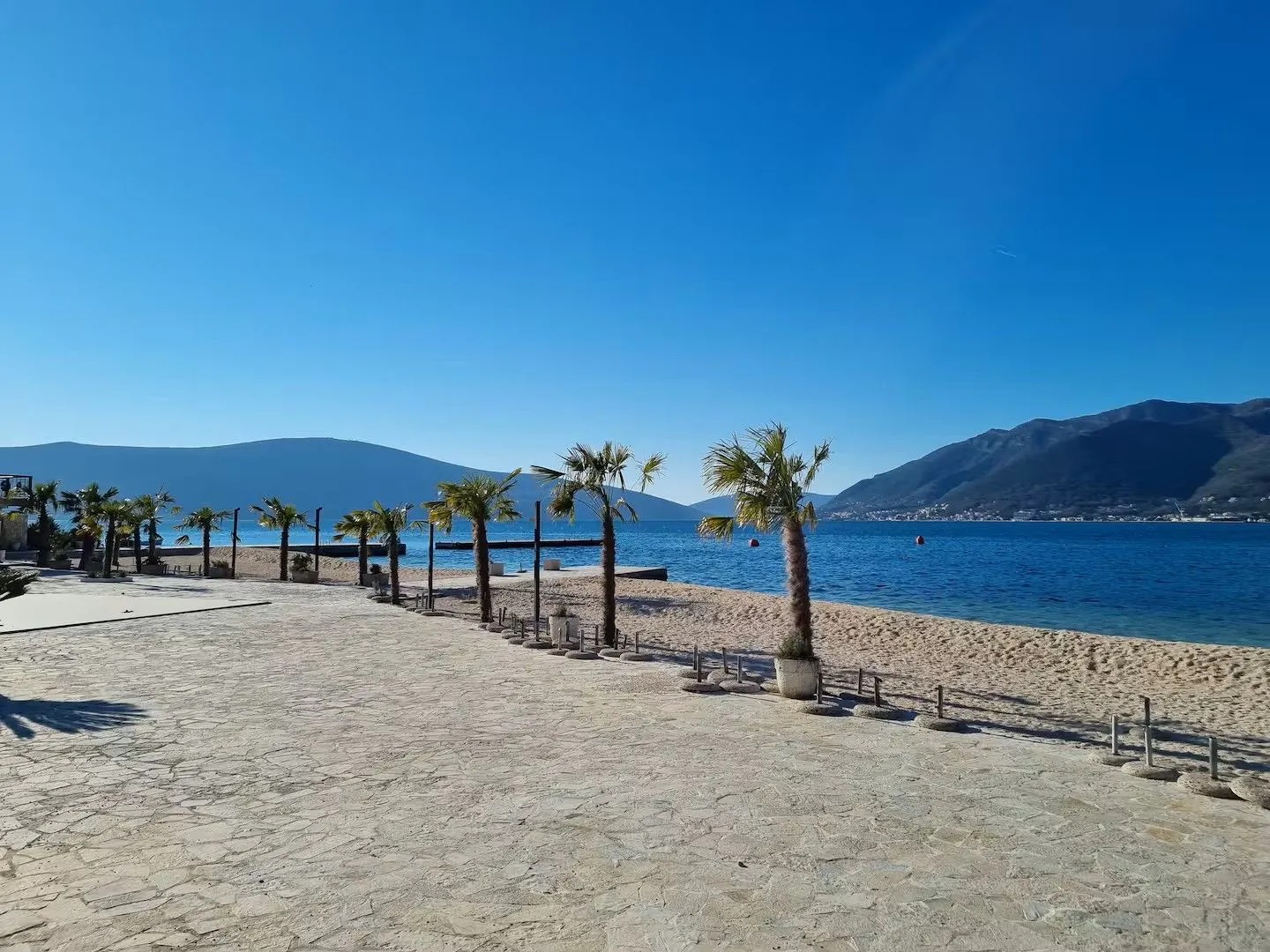 Popločano šetalište pored pješčane plaže sa palmama i pogledom na more.