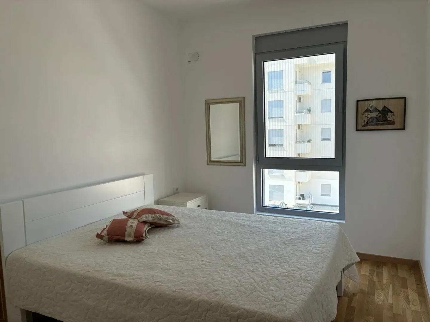 Bright bedroom with a double bed, mirror, and a window with a building view.