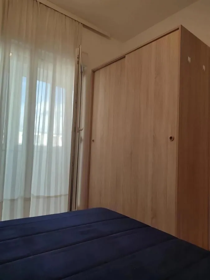 Spavaća soba sa krevetom, drvenim ormarom i balkonskim vratima sa bijelim zavjesama.