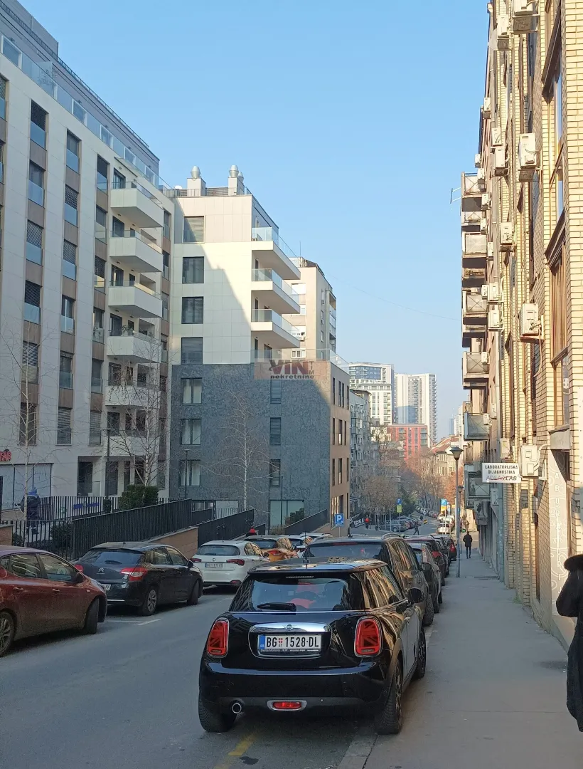 Pogled na ulicu sa parkiranim automobilima i stambenim zgradama.