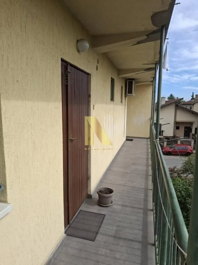 Dugački natkriveni balkon sa metalnom ogradom i ulaznim vratima.