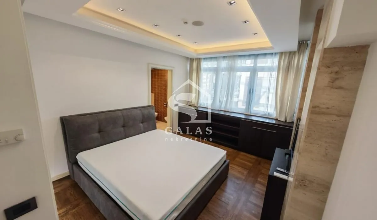 Furnished bedroom with a double bed, parquet flooring, and a dropped ceiling.