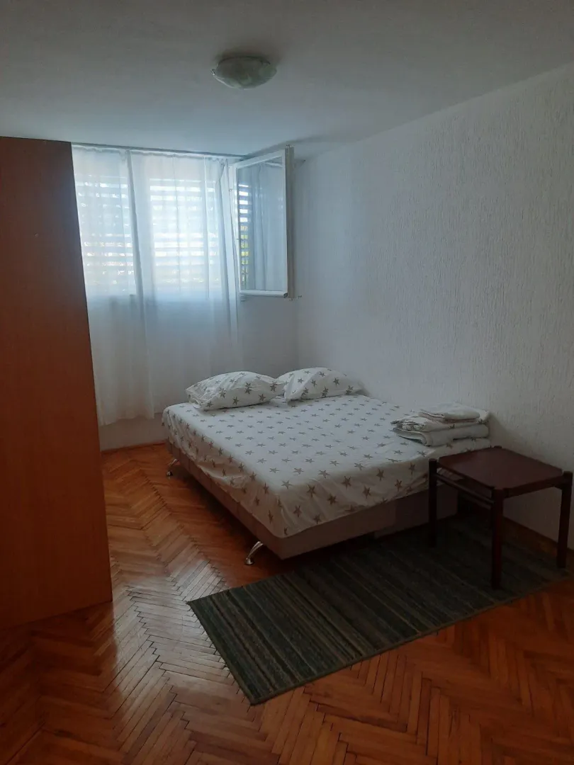 Spavaća soba sa bračnim krevetom, drvenim parketom i prozorom sa bijelim zavjesama.