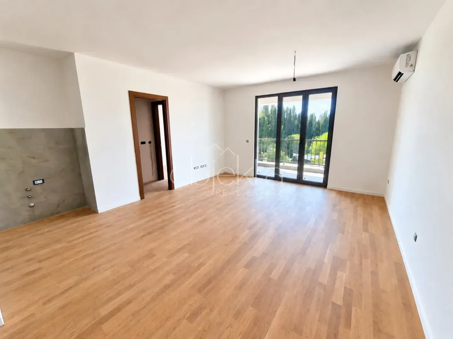 Prazna soba sa drvenim podom, bijelim zidovima i balkonskim vratima.
