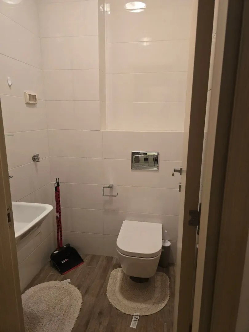 Manje kupatilo sa WC šoljom, umivaonikom i bijelim pločicama.