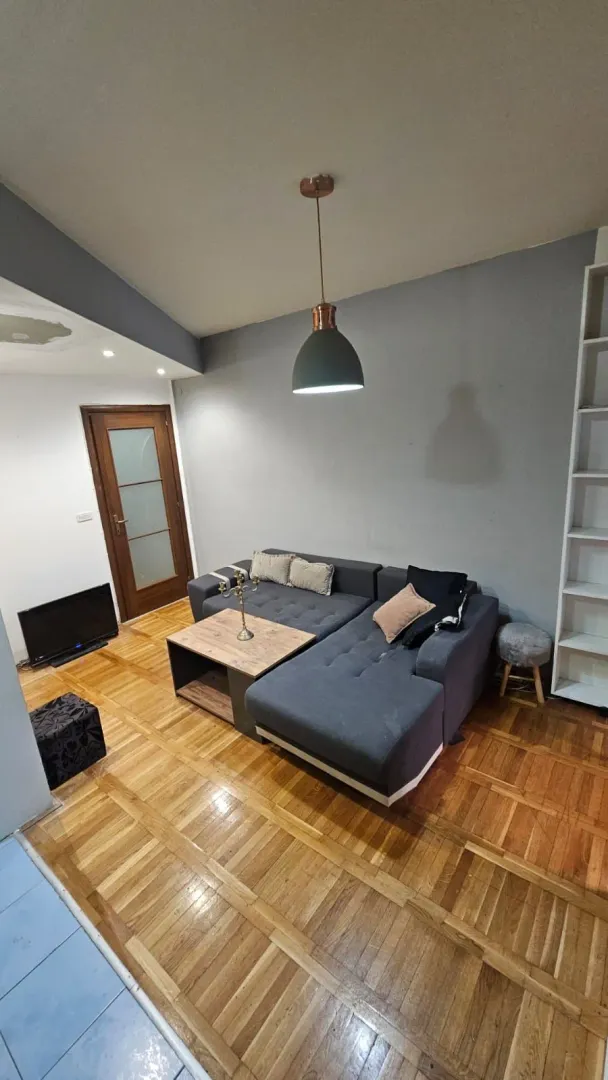 Dnevna soba sa ugaonom garniturom, drvenim podom i visećom lampom.